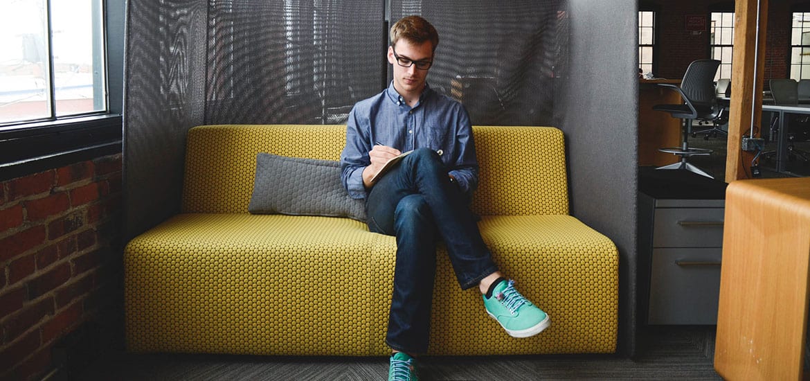 Young man using a tablet on a yellow sofa in a modern office setting.