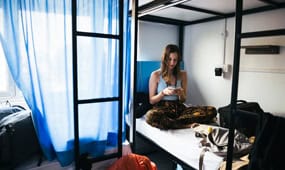 Young woman using her phone in a dorm room with bunk beds and blue curtains.