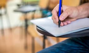 Person writing notes with a pen in a classroom setting.