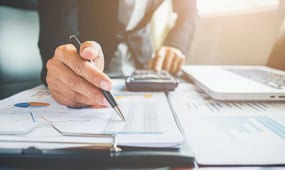 Person signing documents at a desk with tech tools and charts.