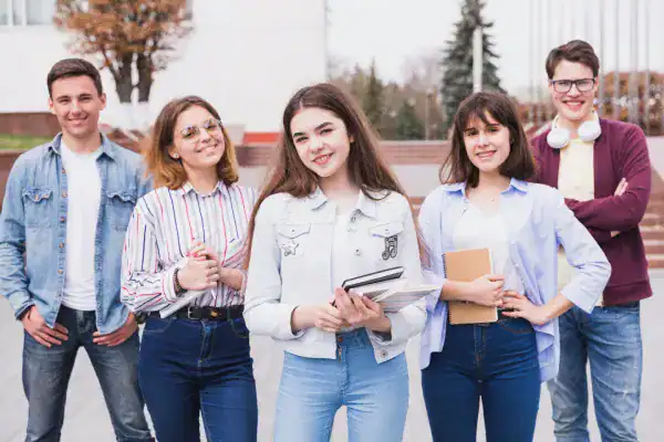Group of diverse students smiling outdoors with books and tablets.