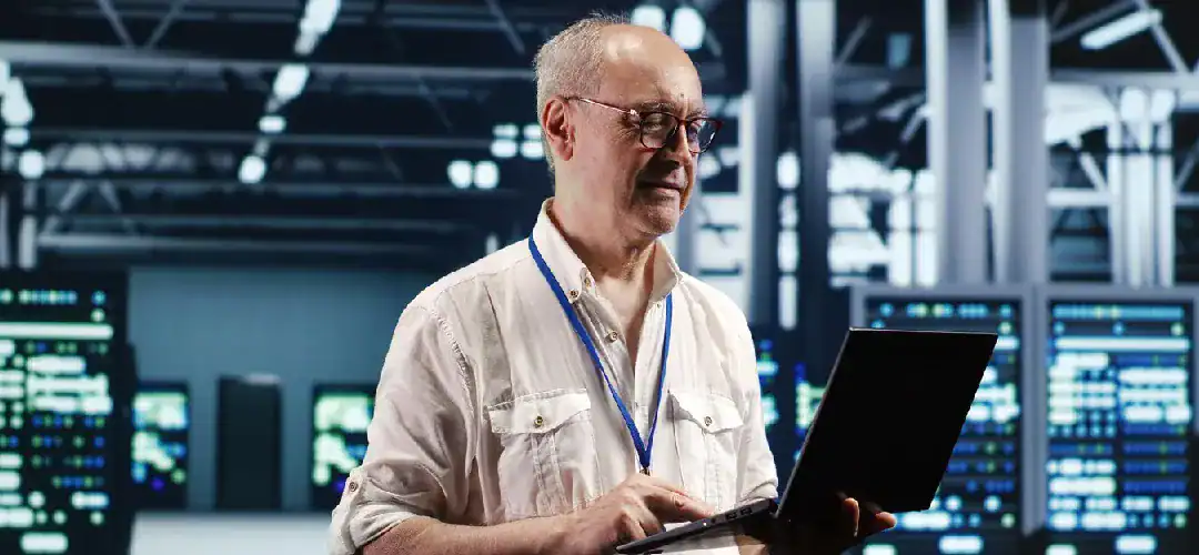 Senior man working on a laptop in a high-tech data center environment.