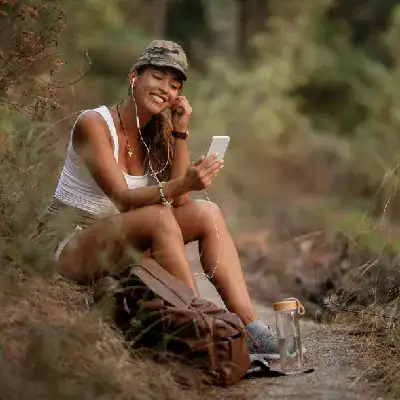 Young woman using smartphone outdoors, smiling, with a backpack and water bottle nearby.