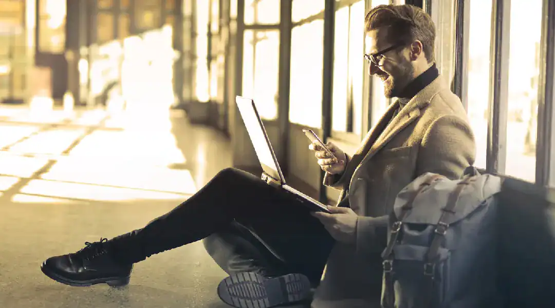 Man using tablet with laptop and backpack in a modern airport setting.
